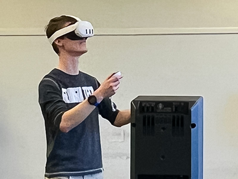 A student wears a VR headset next to a speaker.
